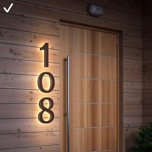 Peut inclure: Numéro de maison éclairé 108 en bronze foncé, monté sur un mur en bois. Les chiffres sont rétro-éclairés d'une lueur chaleureuse, créant une ambiance accueillante. Une porte en bois avec une poignée argentée est à droite.