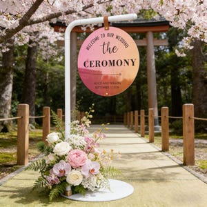 Puede incluir: Un letrero de ceremonia de boda con un degradado rosa y naranja, que muestra "Welcome to our Wedding" y "the Ceremony". El letrero está sostenido por un soporte de metal blanco, con un arreglo floral en la base. También se ven los nombres "Alice and Mason" y la fecha "17 de septiembre de 2026".