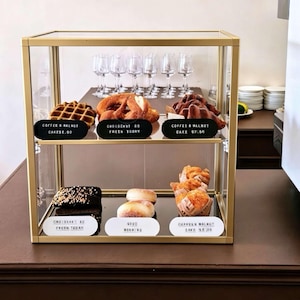 May include: A gold-framed glass display case containing two shelves of pastries. The top shelf holds a waffle, a pretzel, and a cake with labels reading Coffee & Walnut Cake $8.00, Croissant $5 Fresh Today, and Coffee & Walnut Cake $7.50. The bottom shelf holds a chocolate-topped pastry and muffins with labels reading Croissant $5 Fresh Today, Good Morning, and Coffee & Walnut Cake $5.00.