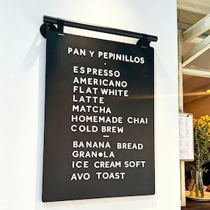 May include: A black rectangular menu board with white lettering, hanging on a white wall. The menu lists coffee drinks like espresso, latte, and matcha, along with food items such as banana bread and avocado toast.