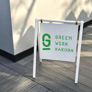 May include: A white A-frame sidewalk sign featuring a square panel suspended by two gold-toned metal clips. The sign displays a large green letter G logo and the green text GREEN WORK HAKUBA in a stencil-style font on a white background.