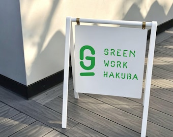 May include: A white A-frame sidewalk sign featuring a square panel suspended by two gold-toned metal clips. The sign displays a large green letter G logo and the green text GREEN WORK HAKUBA in a stencil-style font on a white background.