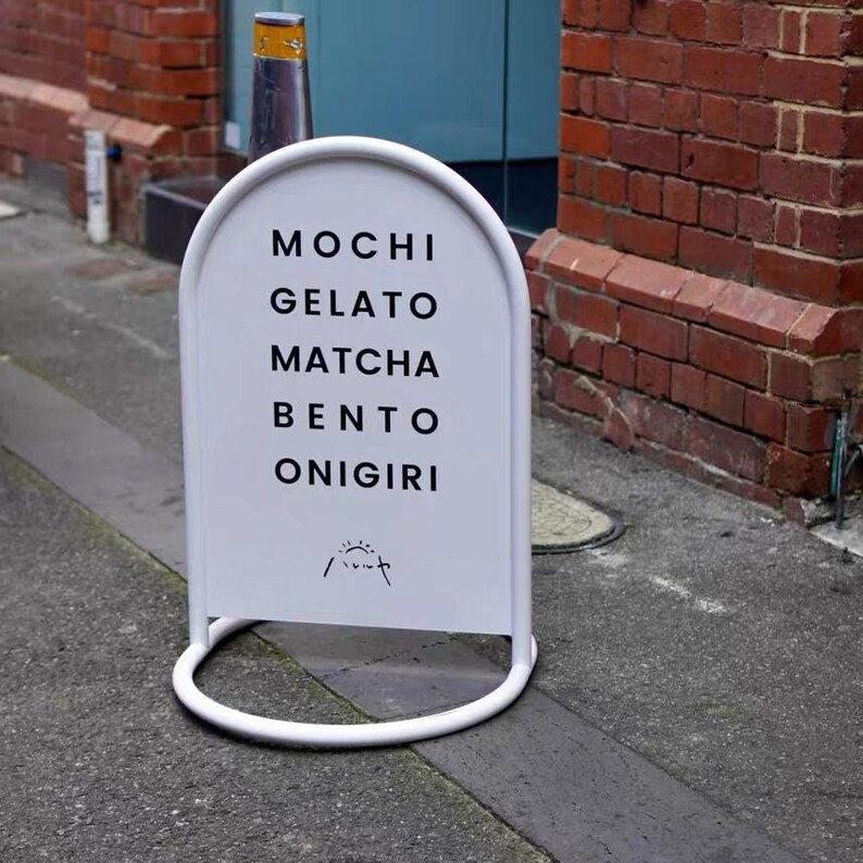 May include: A white, arched sandwich board sign with the words "MOCHI GELATO MATCHA BENTO ONIGIRI" in black. The sign is on a gray sidewalk, with a brick wall and a blue door in the background.