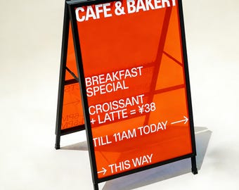 May include: A-frame sign advertising a cafe and bakery. The sign features a black frame and orange translucent panels. White text reads "CAFE & BAKERY", "BREAKFAST SPECIAL", "CROISSANT + LATTE", and "THIS WAY". The sign is angled towards the viewer.
