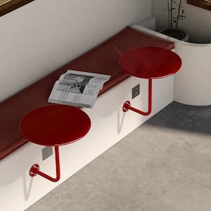 May include: Two red, circular wall-mounted tables with a curved support. The tables are fixed to a white wall, near a burgundy bench with a newspaper. The tables are a modern design, suitable for small spaces.