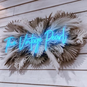 May include: A neon sign with the text "The Vintage Pearl" in blue, surrounded by a wreath of dried pampas grass.