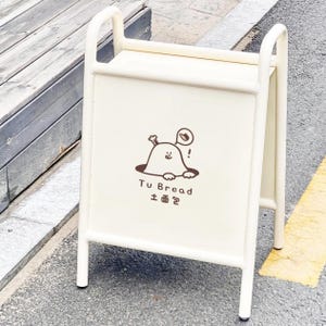 Puede incluir: Un letrero publicitario blanco en forma de caballete con una ilustración de dibujos animados y texto. La ilustración presenta un personaje marrón con un bollo. El texto dice "Tu Bread" en inglés y en chino.