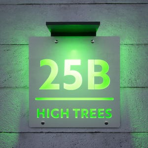 以下が含まれることがあります： 明るい緑色で「25B」と「HIGH TREES」と書かれた、白い背景の照明付き住所看板。看板はコンクリートの壁に取り付けられており、上部に黒いソーラーパネルが付いています。