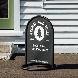 May include: A black metal A-frame sign with rounded top and base. The sign features the text "GEORGE AND WILLY" around a white tree graphic. Below, it reads "GOOD SIGNS FOR GOOD TIMES" and "GEORGEANDWILLY.COM".