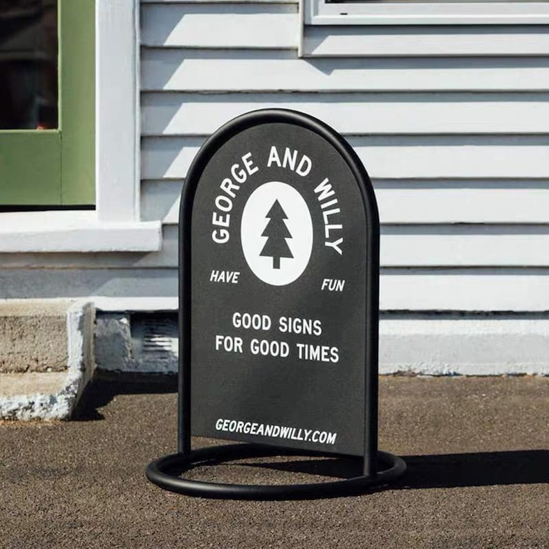 May include: A black metal A-frame sign with rounded top and base. The sign features the text "GEORGE AND WILLY" around a white tree graphic. Below, it reads "GOOD SIGNS FOR GOOD TIMES" and "GEORGEANDWILLY.COM".