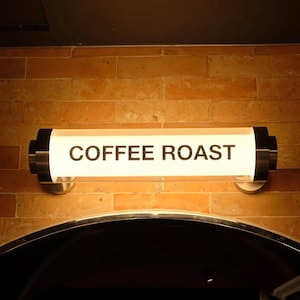 May include: A rectangular, illuminated sign with the words "COFFEE ROAST" in black lettering. The sign is mounted on a brick wall and has a metallic frame with black end caps. The sign is above a dark, arched doorway.