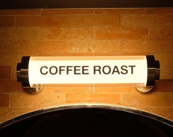 May include: An illuminated sign displaying the words "COFFEE ROAST" in black. The rectangular sign is set against a brick wall and has a metallic frame with black end caps. The sign is positioned above a dark, arched doorway.