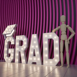 May include: Large white letters spelling "GRAD" with illuminated bulbs, topped with a graduation cap. A wooden artist's mannequin stands next to the "D". The background features a pink and grey striped wall and a light-coloured wooden floor.