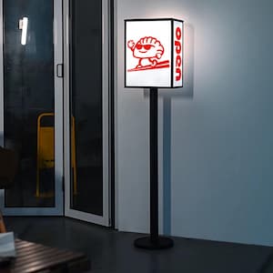 May include: A black, rectangular sign with a cartoon dumpling wearing sunglasses and holding chopsticks. The sign reads "open" in red lettering and is mounted on a black pole. The sign is illuminated.