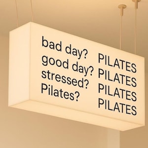 Könnte beinhalten: Ein rechteckiges, beleuchtetes Schild hängt von der Decke. Das Schild zeigt die Sätze „schlechter Tag?“, „guter Tag?“, „gestresst?“ und „Pilates?“ in schwarzer Schrift. Das Wort „PILATES“ wird mehrmals wiederholt.