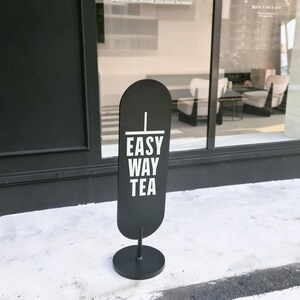 Op de afbeelding: Een zwart, ovaalvormig staand bord met een witte verticale lijn en de tekst "EASY WAY TEA" in witte hoofdletters. Het bord is bevestigd aan een dunne zwarte paal die op een ronde zwarte voet staat, geplaatst op een lichtgekleurde ondergrond.
