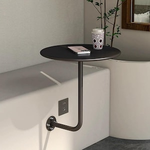Puede incluir: Una mesa auxiliar redonda negra con un solo brazo de soporte fijado a una pared. Un smartphone y un vaso de viaje estampado están sobre la mesa. La mesa está montada en una pared blanca con una toma de corriente.