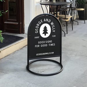 May include: A black metal sandwich board sign with a circular base. The sign has a white tree logo and text that reads "GEORGE AND WILLY HAVE FUN GOOD SIGNS FOR GOOD TIMES GEORGEANDWILLY.COM". The sign is on a concrete surface.