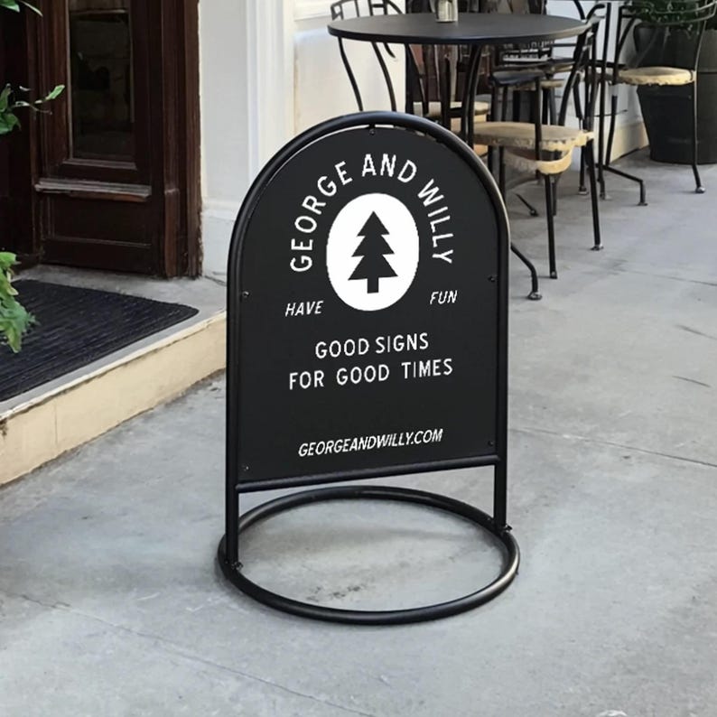 May include: A black metal sandwich board sign with a circular base. The sign has a white tree logo and text that reads "GEORGE AND WILLY HAVE FUN GOOD SIGNS FOR GOOD TIMES GEORGEANDWILLY.COM". The sign is on a concrete surface.