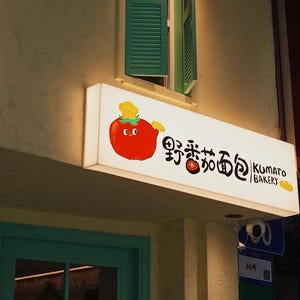 May include: Illuminated bakery sign with a cartoon tomato character holding a rolling pin. The sign reads "野香茄面包" and "KUMATO BAKERY" in black text. The sign is mounted on a beige building exterior, with a green window and door visible.