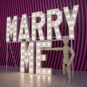 May include: Large white marquee letters spelling "MARRY ME" with illuminated bulbs. A wooden artist's mannequin stands beside the letters, with its arm extended towards the "E". The background features a pink and black striped wall.