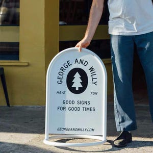 May include: A white, arched-top sign with a rounded base is held by a person. The sign features the text "GEORGE AND WILLY" around a black circle with a tree graphic. Below, it reads "HAVE FUN GOOD SIGNS FOR GOOD TIMES" and "GEORGEANDWILLY.COM".