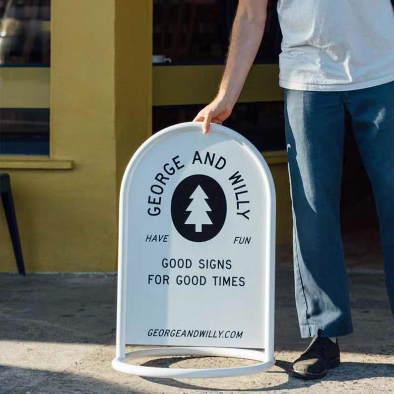 May include: A white, arched-top sign with a rounded base is held by a person. The sign features the text "GEORGE AND WILLY" around a black circle with a tree graphic. Below, it reads "HAVE FUN GOOD SIGNS FOR GOOD TIMES" and "GEORGEANDWILLY.COM".