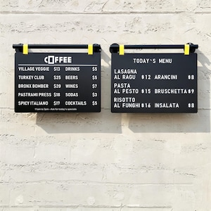 May include: Two rectangular black menu boards with white text mounted on a wall. The left board lists coffee and food items like Village Veggie and Turkey Club. The right board displays Today's Menu with items including Lasagna and Risotto.