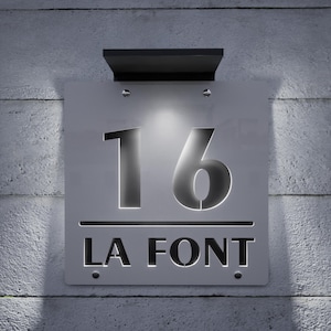 以下が含まれることがあります： スタイリッシュなフォントで「16」の数字と「LA FONT」の名前が表示された、モダンな照明付き住所プレート。銀色のプレートは、テクスチャ加工されたコンクリートの壁に取り付けられており、上部には黒い照明器具があります。