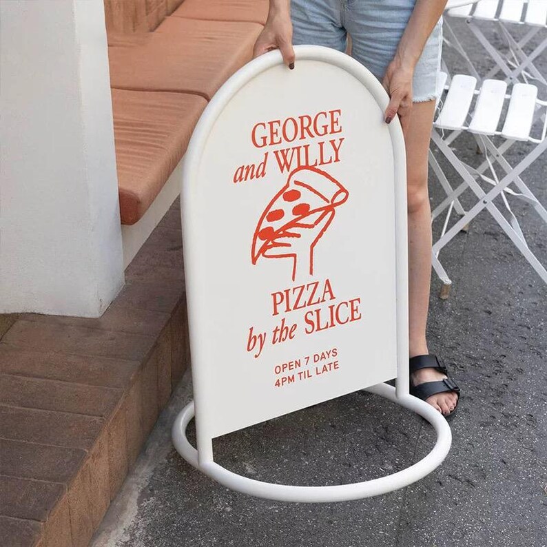 May include: White, arched sidewalk sign with red text and a pizza slice graphic. The sign reads "GEORGE and WILLY PIZZA by the SLICE" and lists the hours of operation. The sign is held upright by a person.