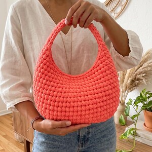 May include: A bright coral-colored handbag crafted with a dense, textured crochet stitch pattern. The bag features a rounded, crescent-like shape with a single integrated handle at the top, showcasing a uniform, knotted yarn construction throughout.