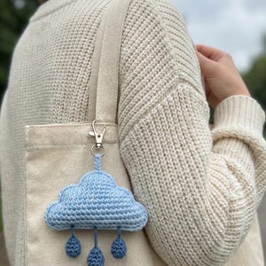 Puede incluir: Un llavero de nube de ganchillo azul claro con tres gotas de lluvia colgantes. La nube está sujeta a un cierre metálico plateado, colgando de un bolso tote beige. El fondo muestra a una persona con un suéter color crema.
