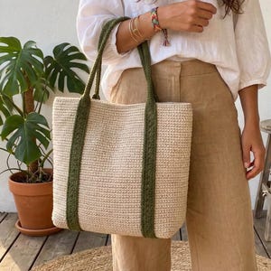 Può includere: Una borsa tote beige con cinghie verde oliva. La borsa ha una trama intrecciata e una forma rettangolare. Le cinghie sono attaccate alla parte superiore della borsa e sono abbastanza lunghe da essere portate a spalla. La borsa è tenuta da una persona.