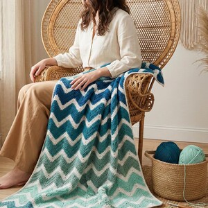 Könnte beinhalten: Eine blau, weiß und türkis gemusterte Decke mit Zickzackmuster, die über einem Korbstuhl drapiert ist. Die Decke besteht aus einem weichen, strukturierten Material. Ein Korb mit zwei Wollknäueln steht in der Nähe. Der Stuhl steht in einem hellen Raum mit einem Fenster.