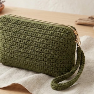 May include: Olive green crochet clutch with a gold-tone zipper and wrist strap. The rectangular bag is made of a textured, hand-stitched yarn. The zipper runs across the top of the bag. The wrist strap is attached to the side of the bag.