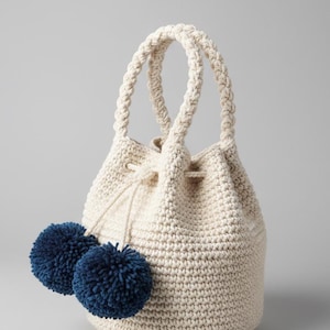 Può includere: Borsa a secchiello all'uncinetto color crema, con manici intrecciati e chiusura con coulisse. La borsa presenta due grandi e soffici pompon blu navy. Un accessorio elegante.