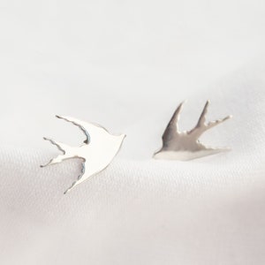 May include: A pair of silver-colored bird-shaped stud earrings. The earrings are displayed on a white, textured fabric. The birds are facing opposite directions, with their wings outstretched.
