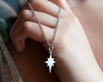 May include: Silver necklace with a starburst pendant. The pendant has a textured, possibly mother-of-pearl, appearance. The chain is a delicate, linked design. The necklace is being held up, showcasing its design and detail.