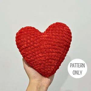 May include: A bright red heart-shaped plush object made of thick, chunky yarn with a visible crocheted texture. A white circular graphic in the lower right corner contains the black text PATTERN ONLY, indicating this is a digital design.