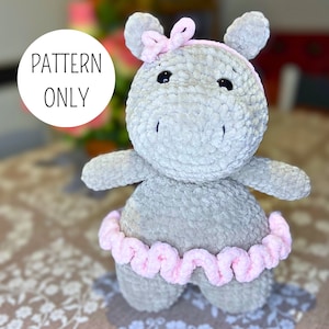 May include: A gray crocheted hippopotamus wearing a pink tutu and a pink bow. The hippopotamus is sitting on a patterned surface.