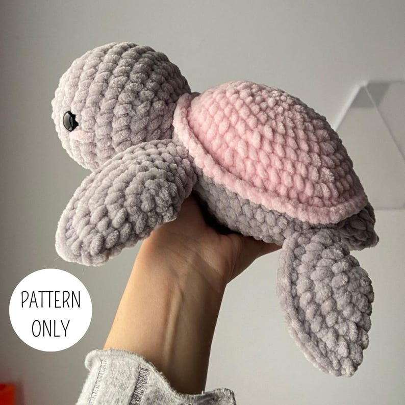 May include: A crochet turtle plush toy made from soft, chunky grey yarn with a light pink shell. The turtle features a rounded head, two front flippers, and two back flippers, with black plastic eyes. A white circular graphic displays the text PATTERN ONLY.