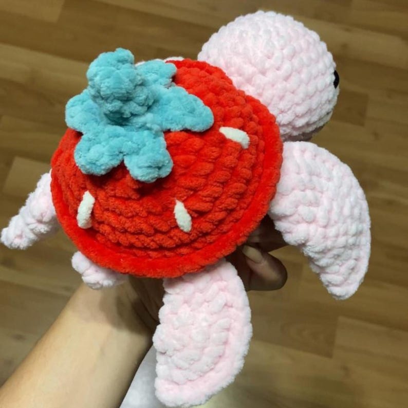 May include: A handmade crochet turtle plushie with a strawberry shell. The turtle's shell is red with white seed details and a blue leafy top. The turtle's head, flippers, and limbs are light pink.