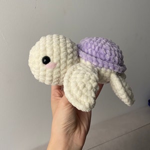 Può includere: Un giocattolo di tartaruga all'uncinetto con un corpo bianco e un guscio viola. La tartaruga ha un occhio nero e un naso rosa.
