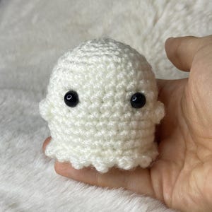 May include: A small, handmade, white crochet ghost with two black safety eyes. The ghost has a rounded body and a ruffled bottom edge. The ghost is a cute, whimsical, and spooky decoration.