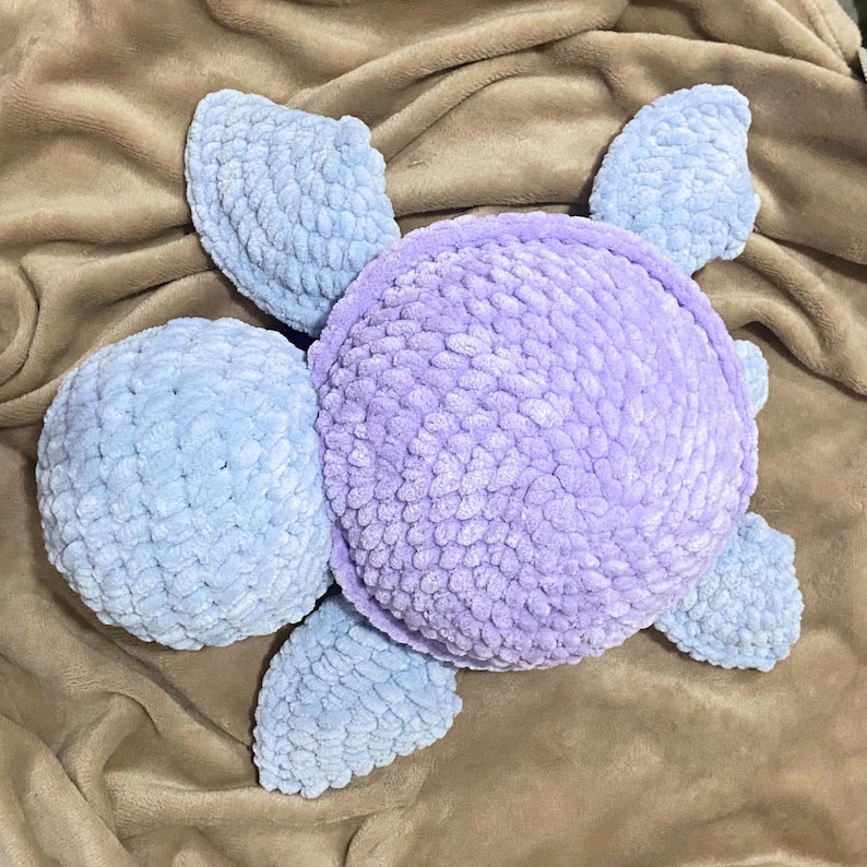 May include: A handmade, plush turtle in shades of lavender and light blue. The turtle's body is a soft, textured material, with a round lavender shell and light blue head and flippers. The turtle is lying on a brown surface.