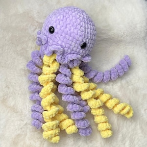 Könnte beinhalten: Ein handgefertigtes Kuscheltier in Form eines Oktopus in Lavendel und Gelb. Der Oktopus hat einen runden Kopf mit schwarzen Knopfaugen und lockigen Tentakeln. Das weiche, strukturierte Garn verleiht dem Spielzeug ein kuscheliges Aussehen, ideal für Kinder oder als Dekoration.