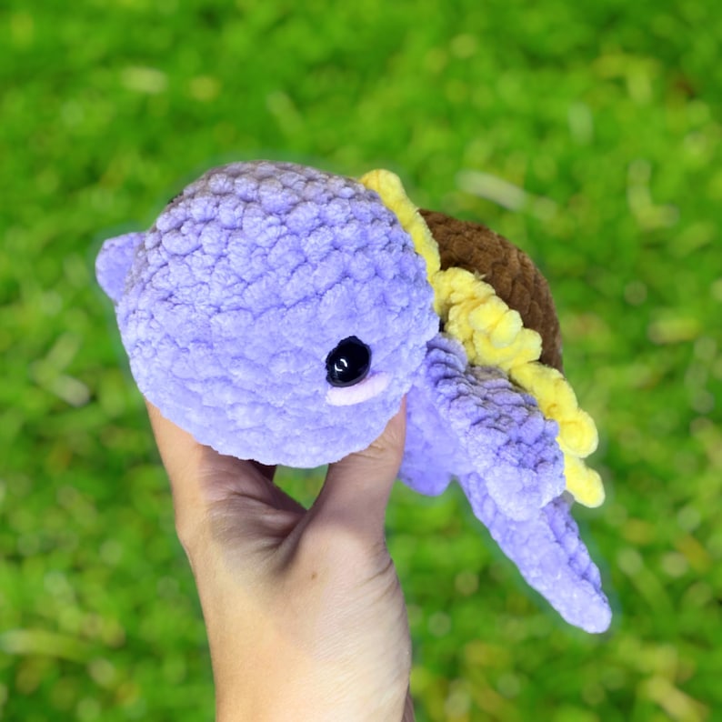 May include: A handmade crochet turtle in shades of lavender, brown, and yellow. The turtle has a textured, plush appearance with black button eyes. The turtle is a soft, cuddly toy.