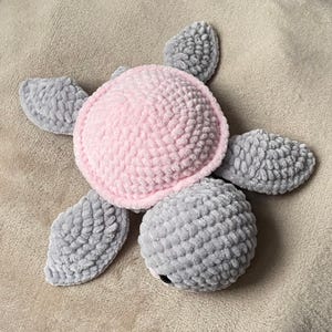 May include: A handmade plush turtle toy with a pink shell and gray flippers and head. The turtle is made of soft, textured yarn and has a cute, round shape. The toy is designed for children and is a cuddly companion.
