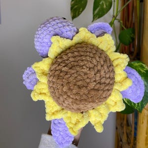 May include: A handmade crochet sunflower plushie. The plushie features a brown center, yellow petals, and lavender accents. The sunflower is made with soft, textured yarn, creating a cozy and whimsical design.