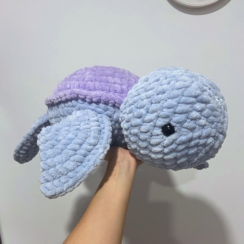 May include: A handmade plush turtle in shades of blue and purple. The turtle has a rounded head and shell, with flipper-like appendages. The eyes are small and black. The soft, textured material gives it a cuddly appearance.